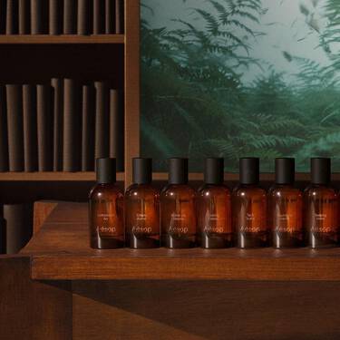 Row of amber glass fragrance bottles with black caps on stepped wooden shelving, with books and fern imagery in the background.