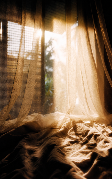 Sheer draped curtains over rumpled fabric near a sunlit window, showing warm beige tones, soft shadows, and translucent folds.