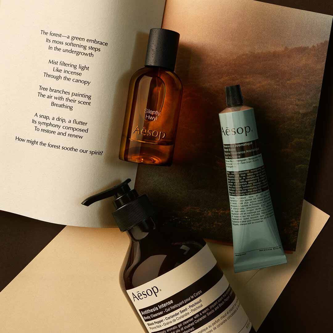 Aesop Silently, Hwyl fragrance bottle, large pump bottle and cosmetic tube on an open book with a woodland photograph.