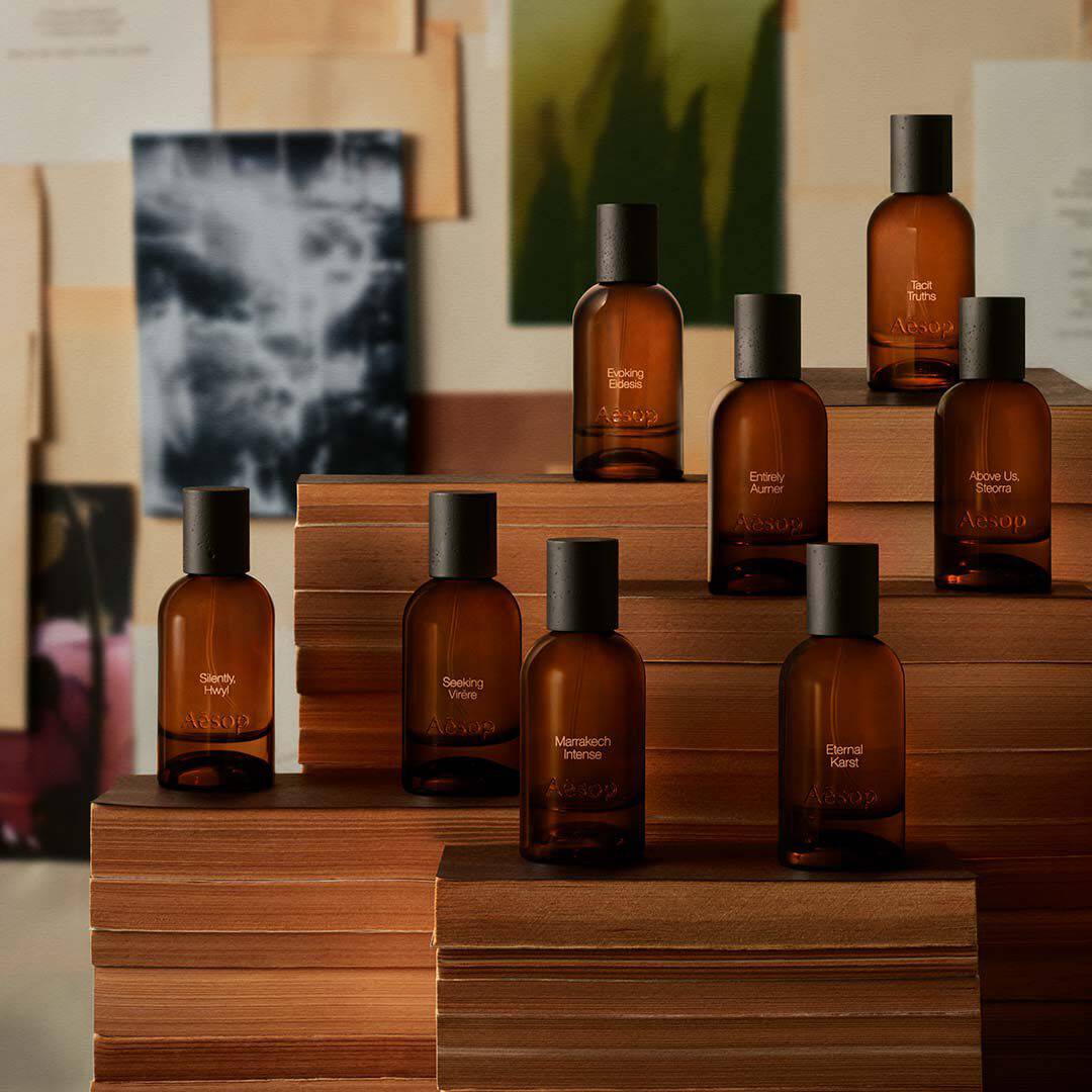 Aesop fragrance bottles arranged on tiered wooden display platforms against an editorial imagery backdrop of prints and paintings.