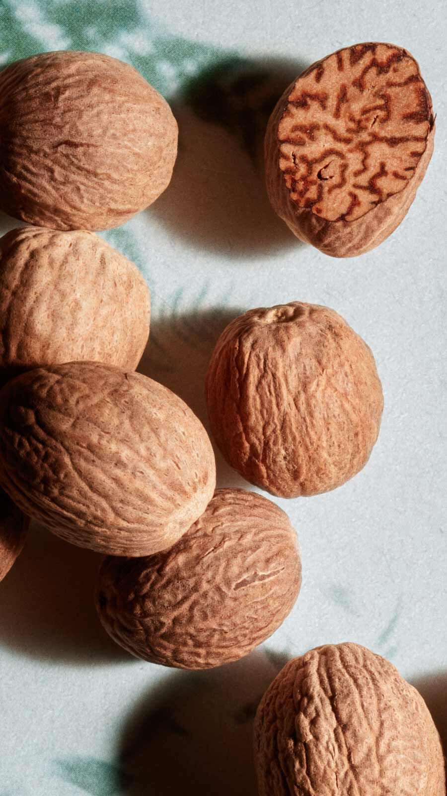 Whole nutmegs scattered on a pale surface, including one cut open to reveal the textured interior.