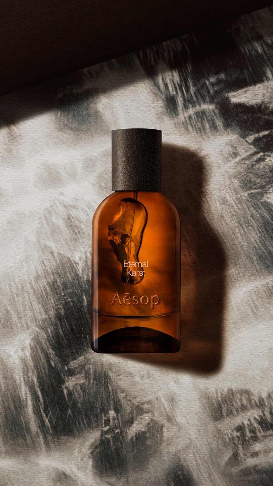 Aesop Eternal, Karst Eau de Parfum bottle with black cap against a pale textured rocky surface with dappled light.