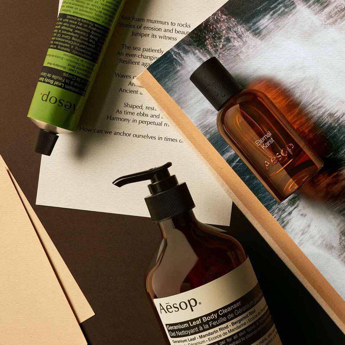 Aesop Eternal, Karst fragrance bottle, large amber pump bottle and cream tube on a dark surface with printed pages.