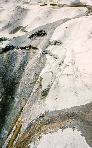 Pale stone and mineral surface with white, grey and charcoal striations, layered textures, and a diagonal fissure crossing the frame.