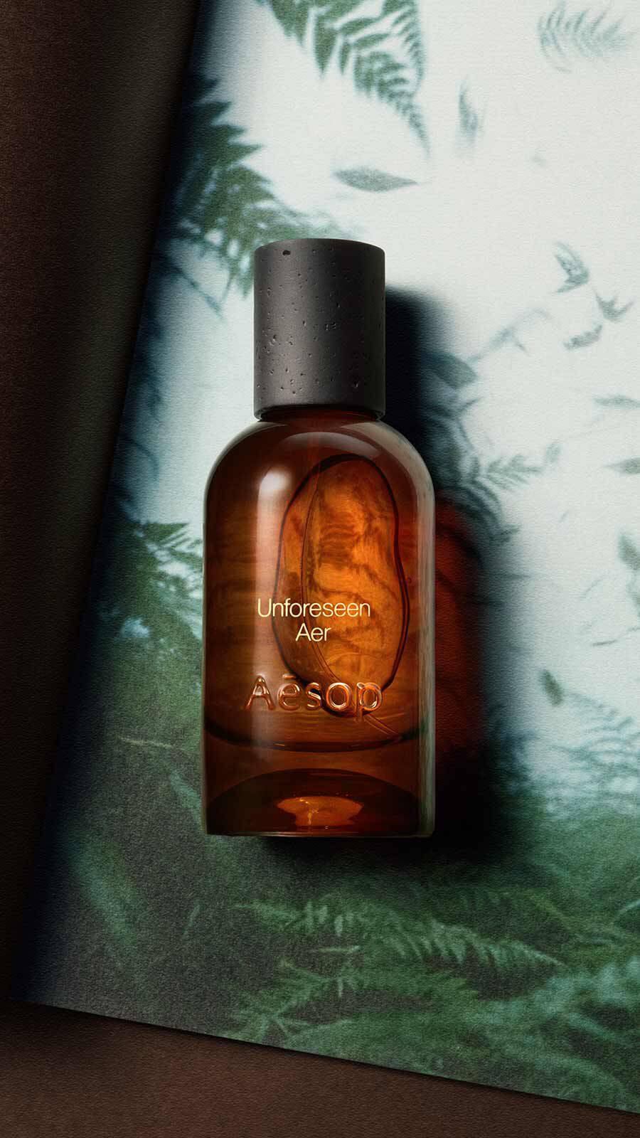 Aesop Unforeseen, Aer Eau de Parfum bottle with black cap against a dark green fern backdrop with soft mottled light.