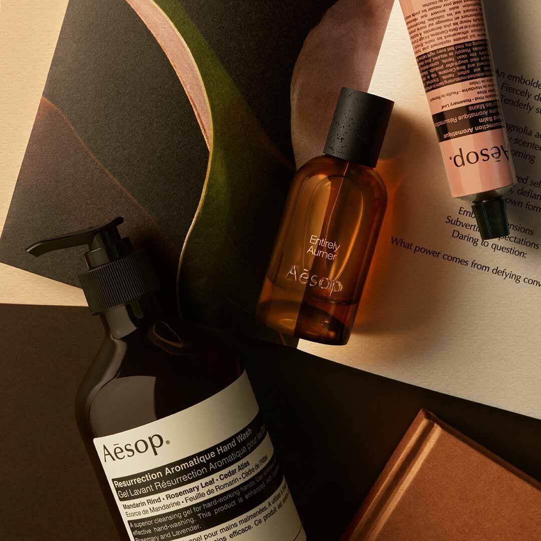 Aesop Entirely, Aurner fragrance bottle, large pump bottle and cream tube on a surface with printed material.