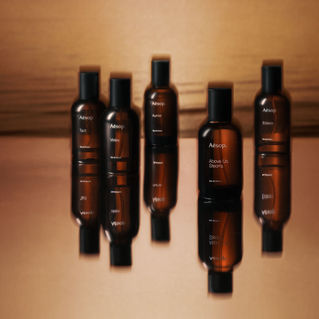 Horizontal arrangement of amber fragrance bottles against a warm brown gradient background, with soft shadows and reflective glass surfaces.