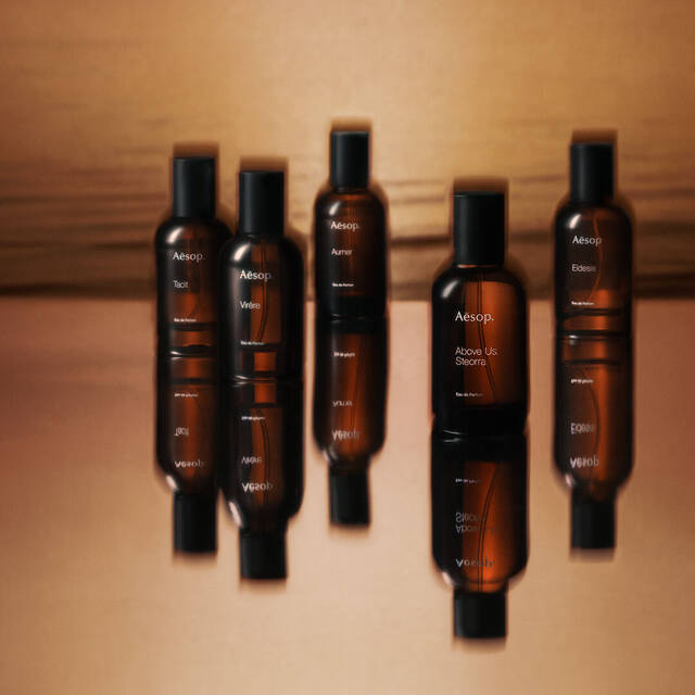 Horizontal arrangement of amber fragrance bottles against a warm brown gradient background, with soft shadows and reflective glass surfaces.
