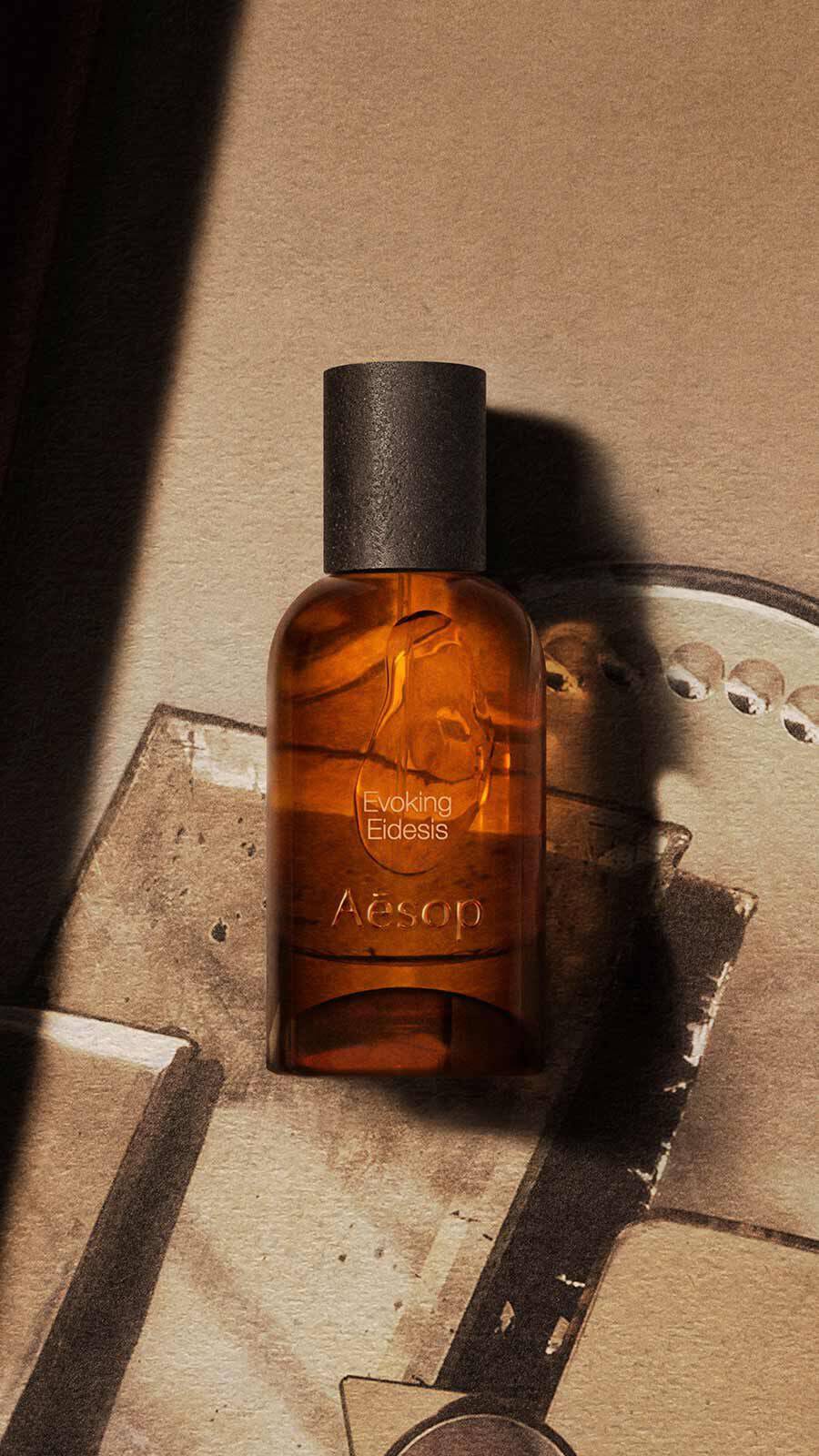 Aesop Evoking, Eidesis Eau de Parfum bottle with black cap on a pale surface beside a decorative metal perforated tray in warm light.