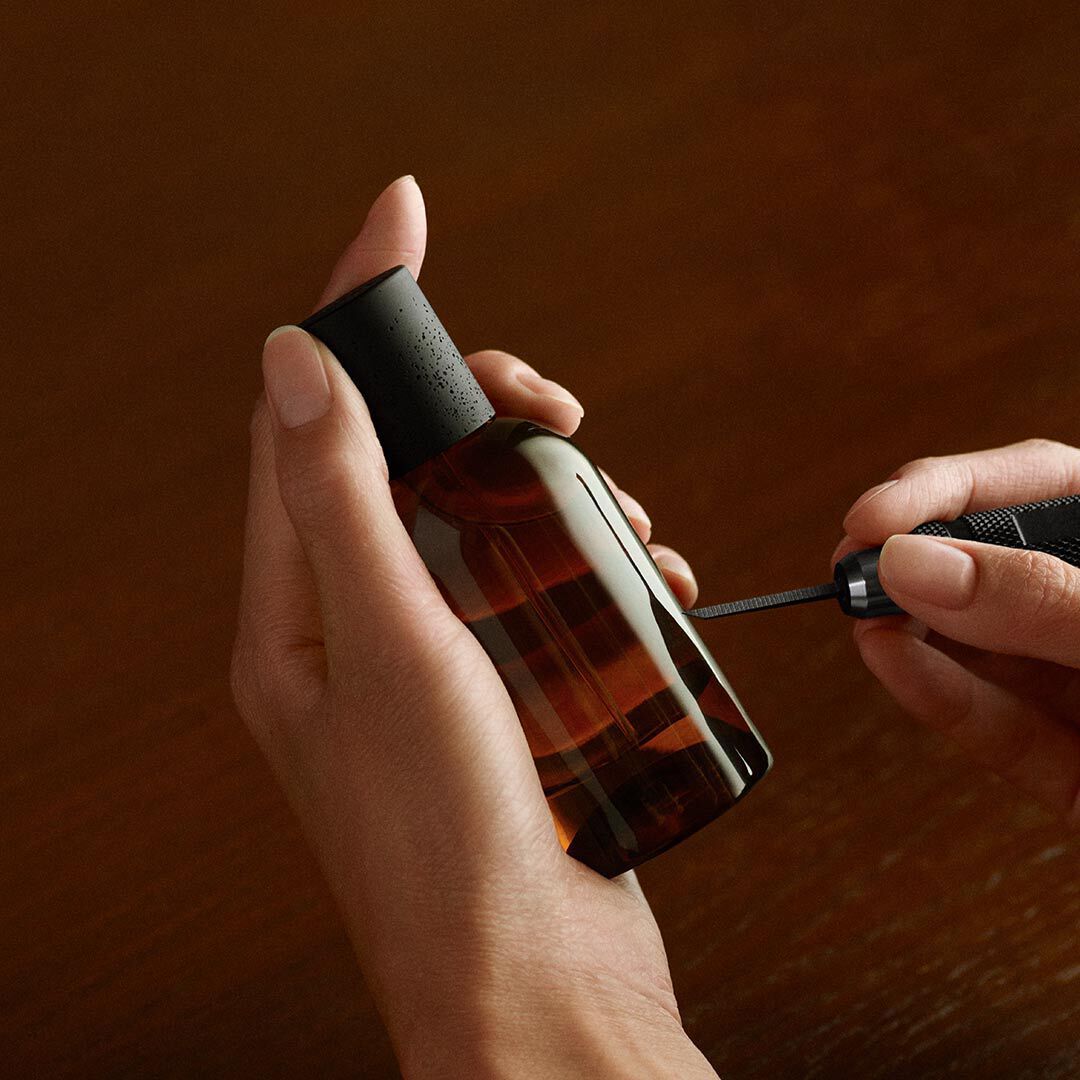 Single amber Aesop bottle lit by a warm glow, framed by curved leaf-like forms in dark green, cream and black tones.