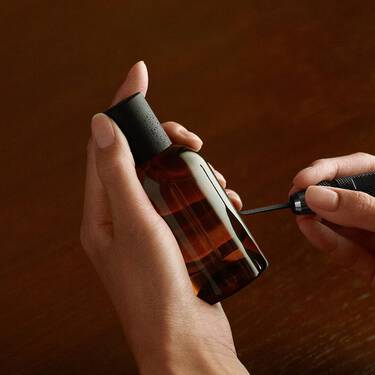 Single amber Aesop bottle lit by a warm glow, framed by curved leaf-like forms in dark green, cream and black tones.