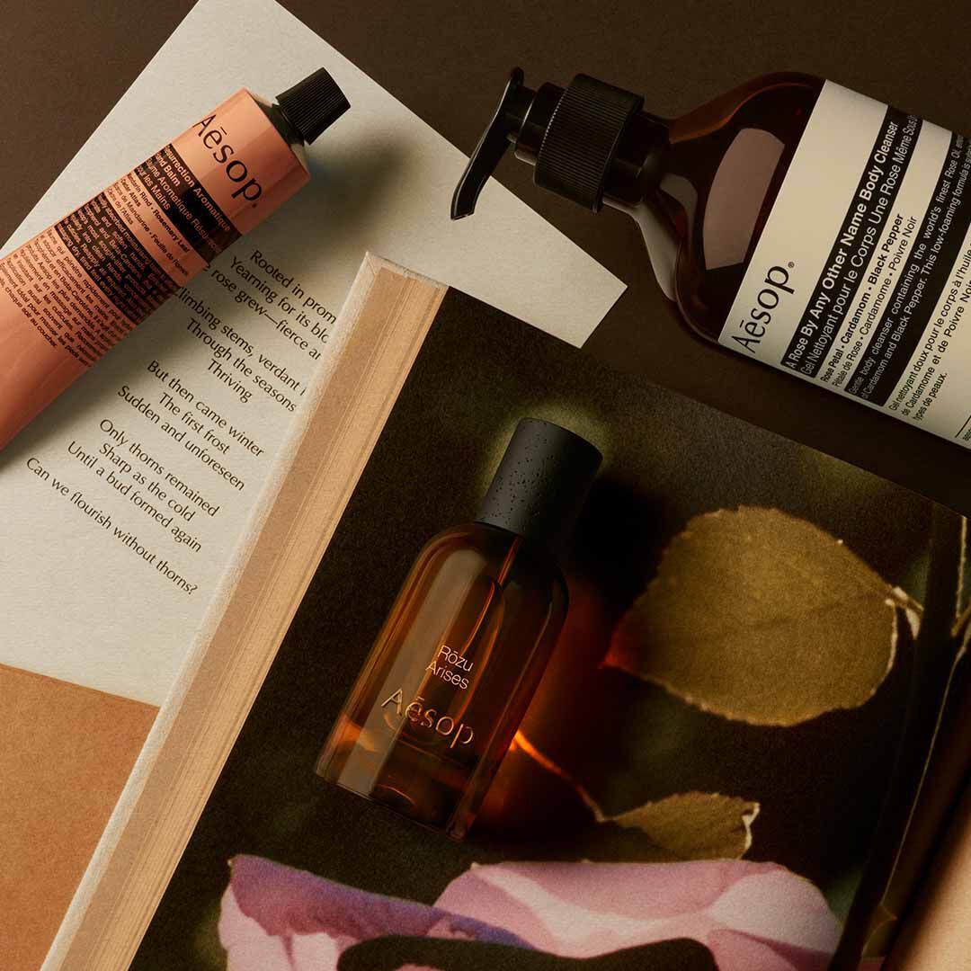 Aesop Rozu Arises fragrance bottle, large pump bottle and mist spray bottle on an open book with a pink rose petal.