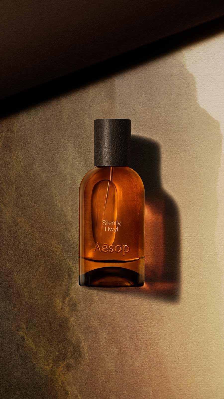 Aesop Silently, Hwyl Eau de Parfum bottle with black cap against a warm golden-brown stone-textured wall with soft light.